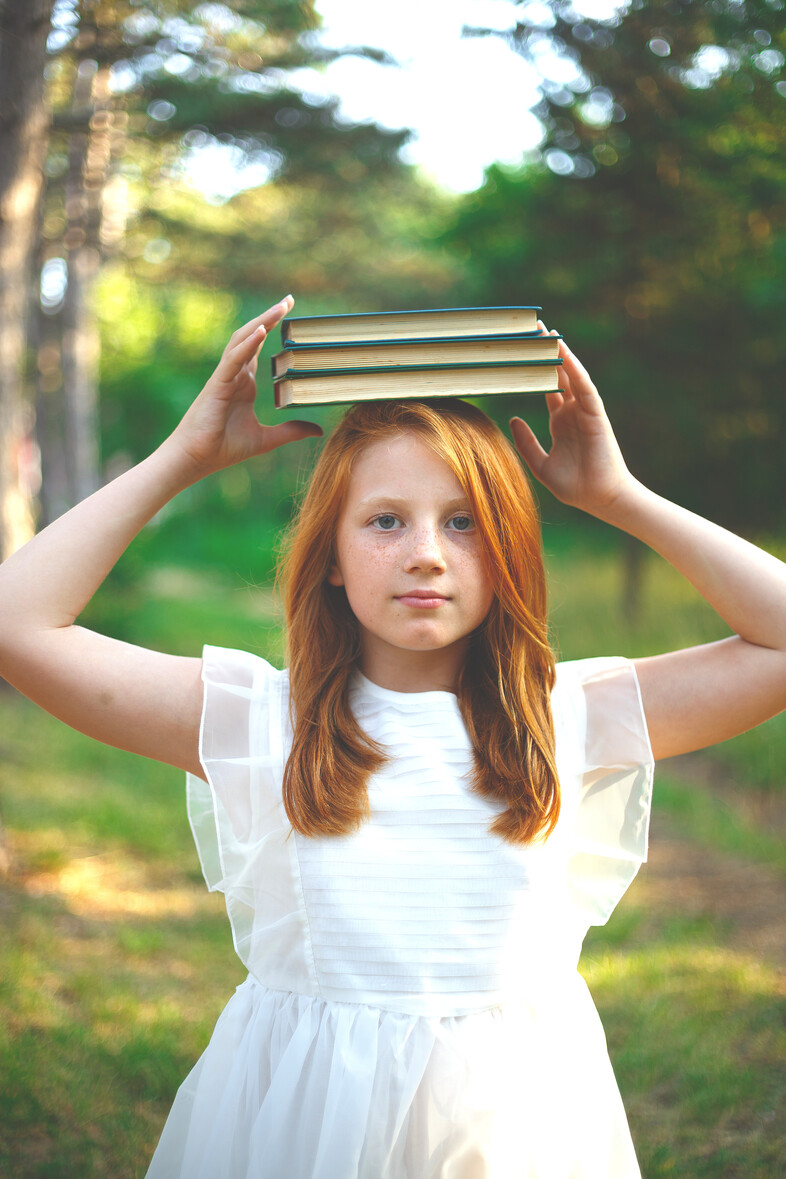 Flicka i vit klänning balanserar tre böcker på huvudet utomhus i en grön skogsmiljö.
