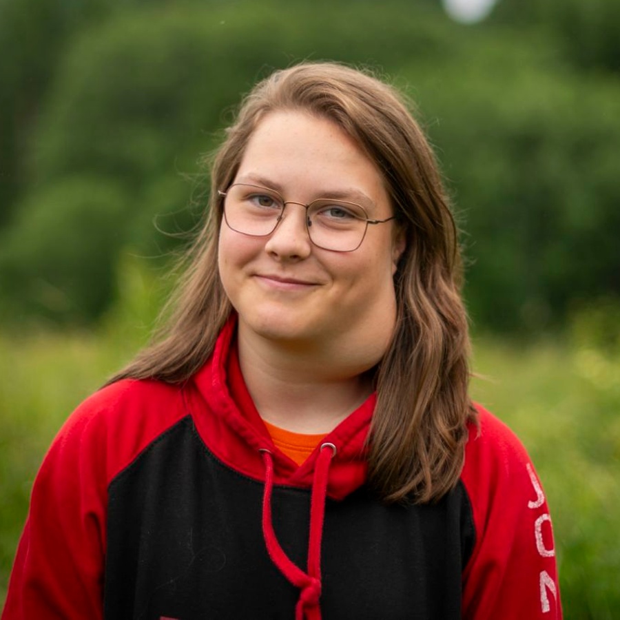 Ung person med glasögon och röd-svart hoodie står utomhus framför en grönskande skog.