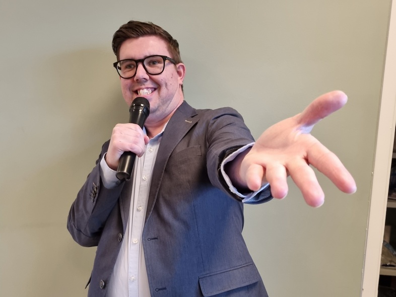 Person i kavaj håller mikrofon och sträcker ut handen mot kameran, ansiktet är suddat.