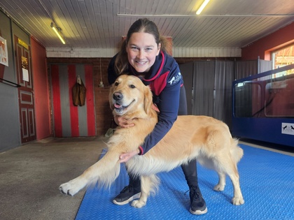 En kvinna ler och håller en glad golden retriever på ett blått golv i ett inomhusutrymme.
