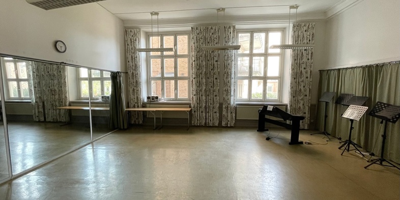 Ljust övningsrum med stora fönster, spegelvägg, elpiano, notställ och bord.