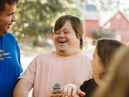 Tre personer står utomhus och skrattar tillsammans, en håller i en mikrofon och en annan i en gitarr.