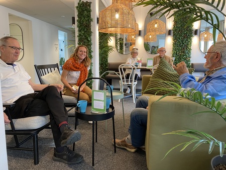 Fyra äldre personer och en yngre kvinna sitter och samtalar i ett ljust, modernt vardagsrum med gröna växter.