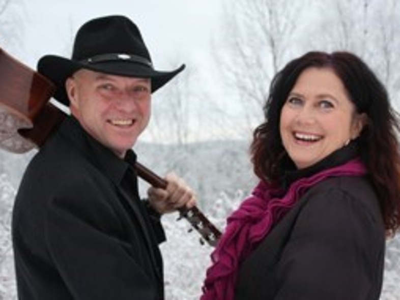 En man med cowboyhatt och gitarr samt en kvinna ler mot kameran i ett vinterlandskap.