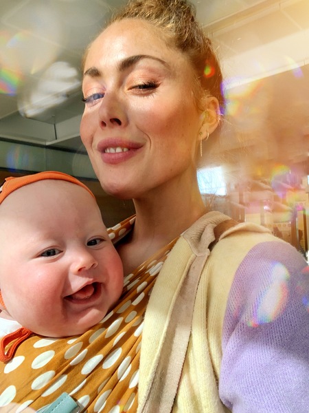 En kvinna tar en selfie med ett leende spädbarn i bärsele, båda ser glada ut i mjukt solljus.