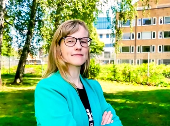 Person står utomhus och tittar in i kameran. Hen har glasögon och längre hår med snedlugg. Solen skiner och det är grönskande bakgrund.