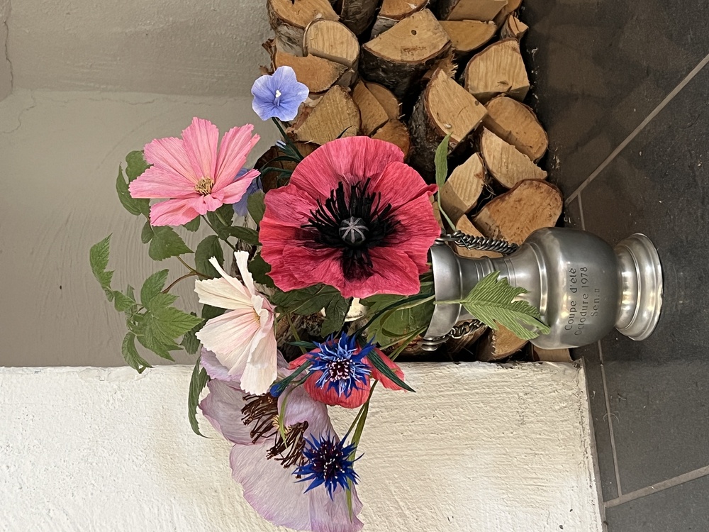 En metallvas med färgglada blommor står på ett golv bredvid en stapel vedträn mot en vit vägg.