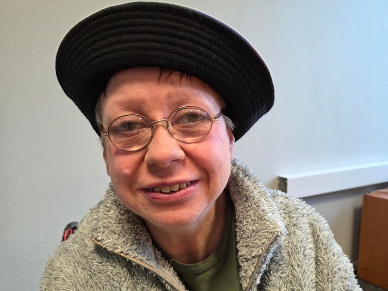 Person med grå fluffig jacka och svart hatt sitter inomhus, ansiktet är suddat.