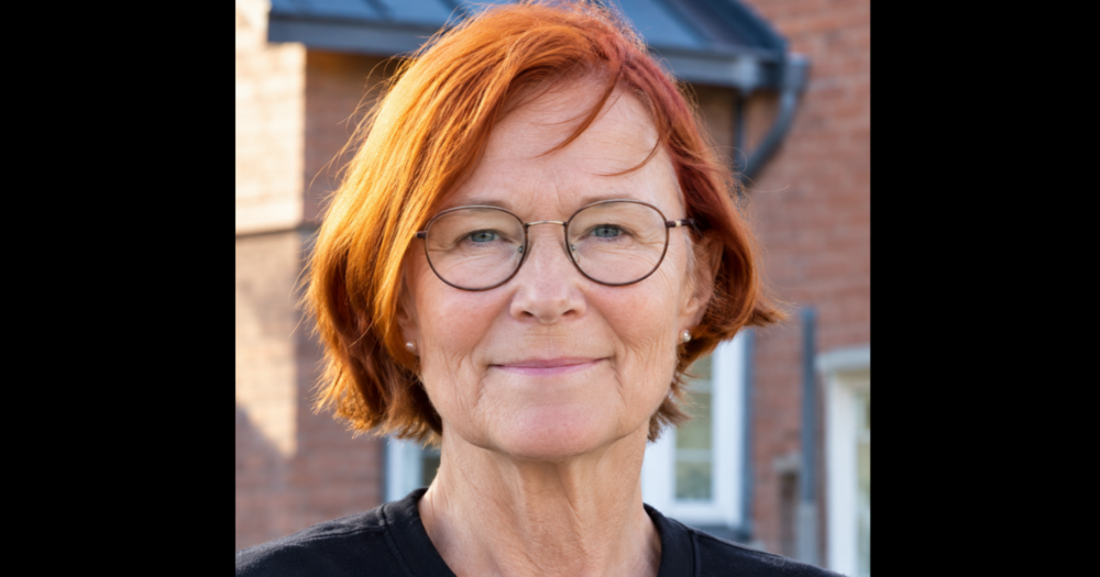 Kvinna med kort rött hår och glasögon står utomhus framför en tegelbyggnad.