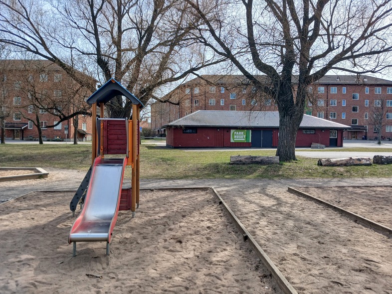 En lekplats med en rutschkana i sandlåda, träd och röda byggnader i bakgrunden.