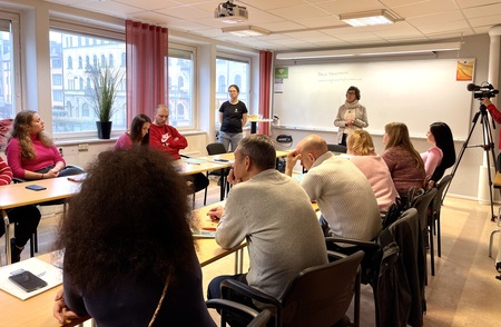 En grupp vuxna sitter runt ett bord i ett klassrum och lyssnar på en presentation vid whiteboard.