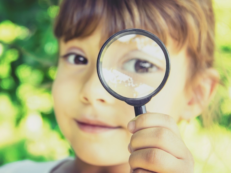 Ett barn håller upp ett förstoringsglas framför sitt öga utomhus, med grönskande bakgrund.
