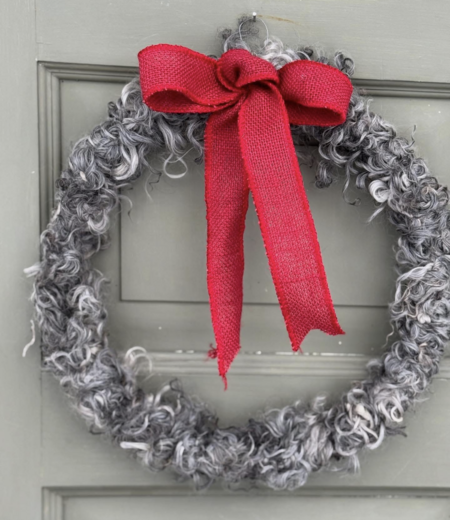 En dörrkrans av lockigt grått material med en stor röd rosett, hänger på en grå dörr.