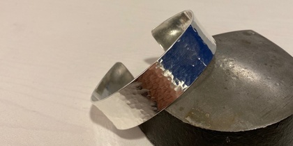 Ett silverfärgat, hamrat armband ligger lutat mot en metallklump på ett ljust underlag.