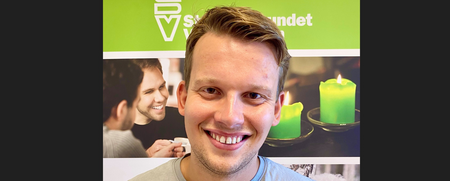 Ung man med ljust hår ler framför en grön skylt och tända ljus i bakgrunden.