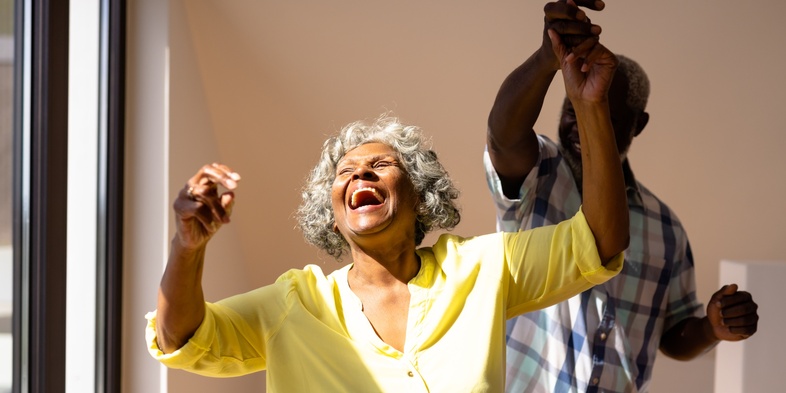 Äldre kvinna i gul skjorta skrattar och dansar med en man i ett soligt rum.