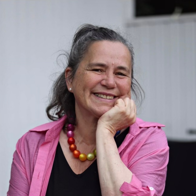 Rinella Rioda  med långt mörkt hår, rosa skjorta och färgglad pärlhalsband sitter med handen mot kinden.