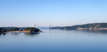 Utsikt över en lugn fjord med en bro i bakgrunden och skogsklädda kullar på sidorna.