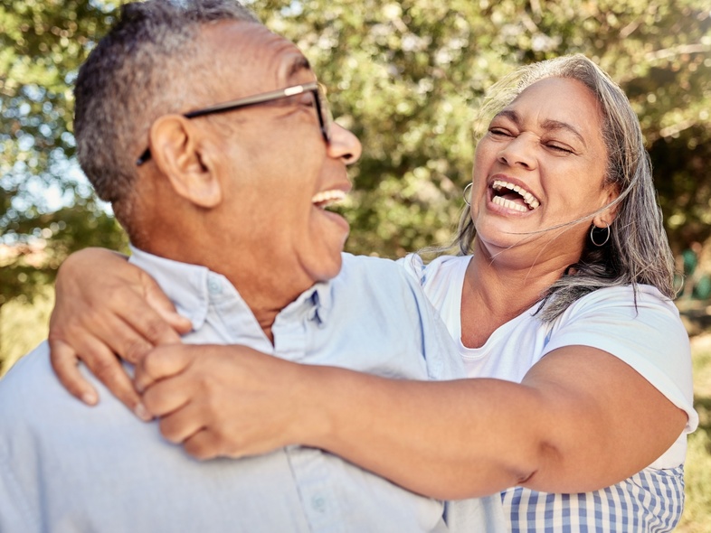 Ett äldre par skrattar och kramas utomhus i solskenet.