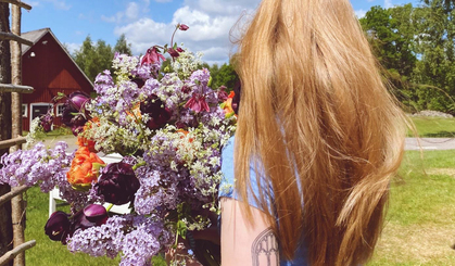 Person med långt blont hår håller en stor bukett blommor utomhus på landet, ryggen vänd mot kameran.