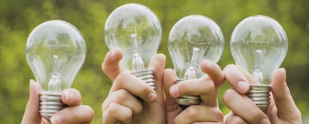 Fyra händer håller upp varsin glödlampa utomhus med grönskande bakgrund.