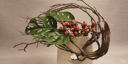 En modern blomsterarrangemang i vit vas med gröna blad, röda bär och böjda grenar mot ljus bakgrund.