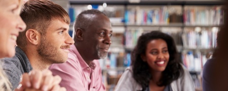 Fyra vuxna personer sitter vid ett bord i ett bibliotek och arbetar tillsammans med datorer och anteckningar.