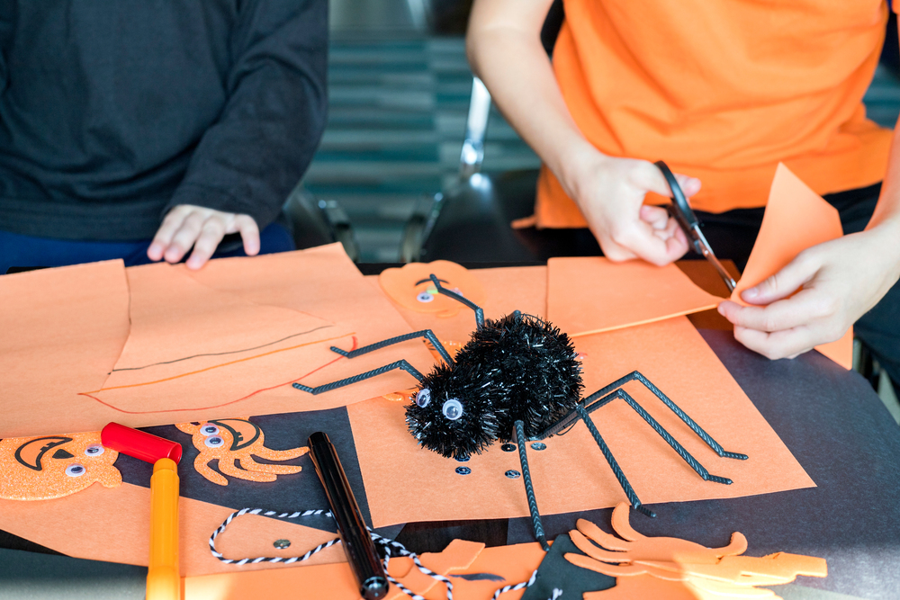 Två personer gör Halloween-pyssel med orange papper, sax och spindeldekoration på ett bord.