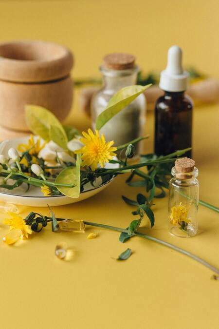 Naturliga ingredienser, blommor och små flaskor på ett gult underlag, med mortel och droppflaska i bakgrunden.