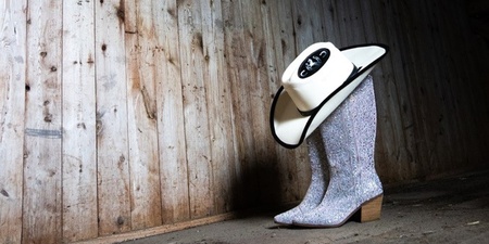 Ett par glittriga cowboyboots med en cowboyhatt ovanpå står på ett trägolv i en gammal lada.