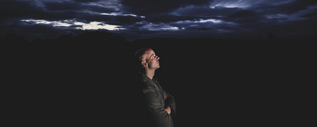 En man står i mörkret och tittar upp mot en dramatisk, molnig himmel i skymningen.