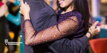 Två personer dansar nära varandra på en social tillställning, leende och festlig stämning.