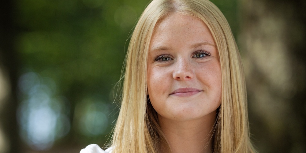 Person med långt blont hår och vit t-shirt står utomhus i en skogsmiljö.