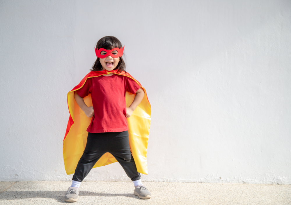 Ett barn utklätt till superhjälte med röd mask och gul cape poserar glatt mot en vit vägg.