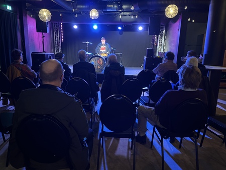 En trumslagare uppträder på en liten scen inför en sittande publik i en mörk konsertlokal.