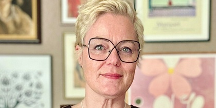 Person med kort blont hår i svart klänning står framför en vägg med tavlor, visar tatuering och smycken.