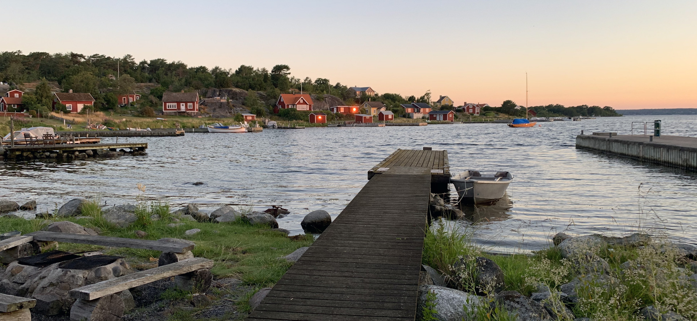 Brygga vid en sjö med små röda stugor längs stranden i solnedgång, båtar i vattnet.