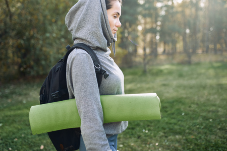 Person i grå hoodie med ryggsäck och yogamatta går utomhus i en park.