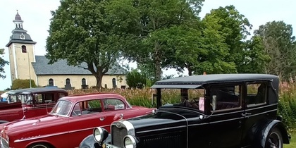 Flera klassiska bilar står parkerade på en gräsmatta med en kyrka och träd i bakgrunden.