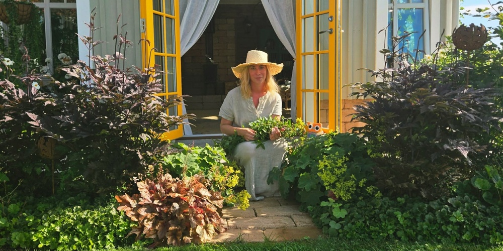 Person med solhatt sitter på trappa vid gul dörr, omgiven av grönskande trädgård och blommor.