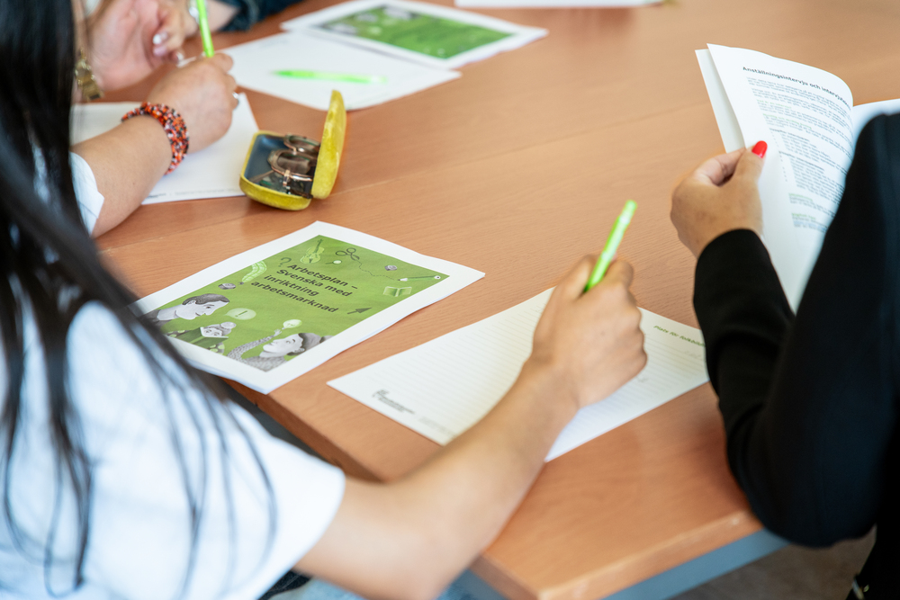 Personer sitter runt ett bord och skriver på papper med gröna pennor under ett möte eller en workshop.