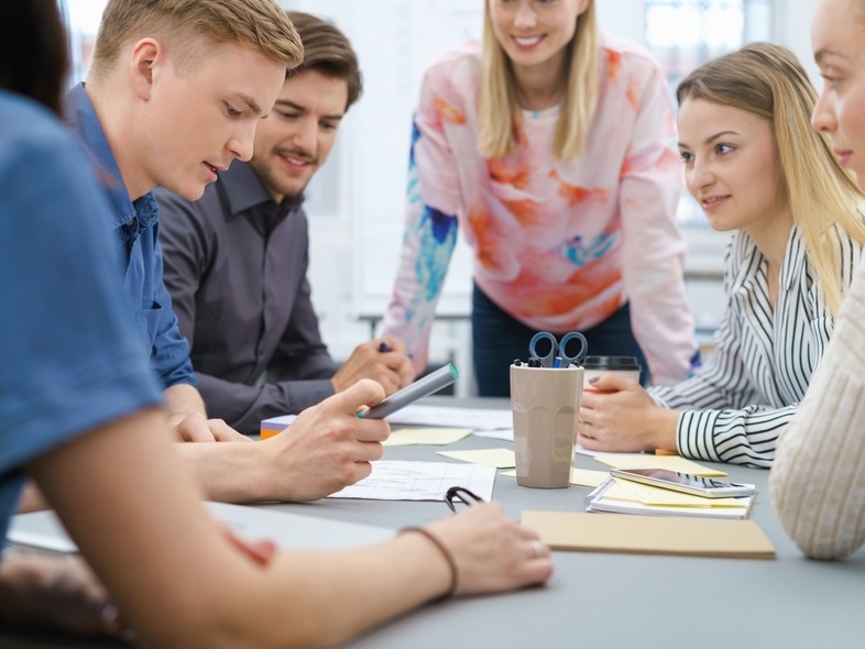 En grupp unga vuxna sitter runt ett bord och diskuterar dokument och en surfplatta.