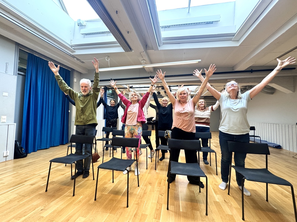 En grupp äldre personer står bakom stolar i ett ljust rum och sträcker armarna uppåt, som i en gruppövning.