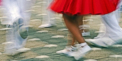 Personer dansar i rörelse, klädda i vita kläder och röd kjol, på en stenlagd gata.
