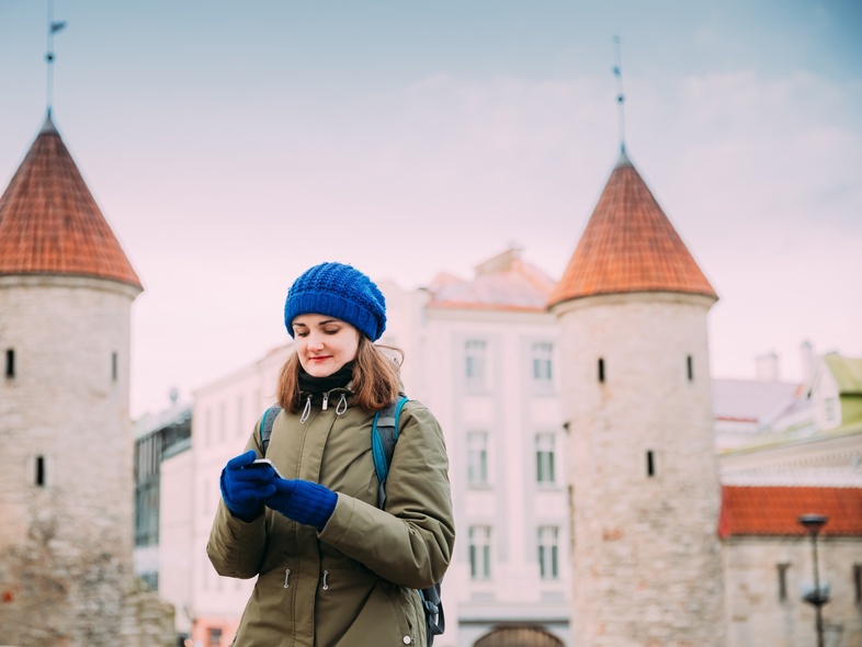 Ung kvinna med blå mössa och vantar står framför medeltida torn och tittar på sin mobiltelefon.