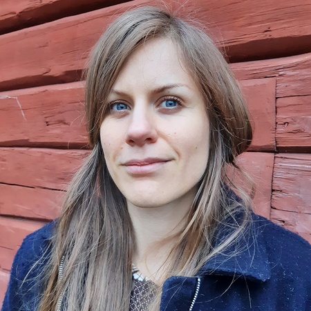 Kvinna med långt blont hår står framför en röd trävägg och tittar in i kameran.