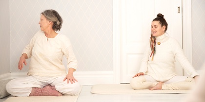 Två personer sitter på yogamattor i ett ljust rum och gör en sittande vridning.