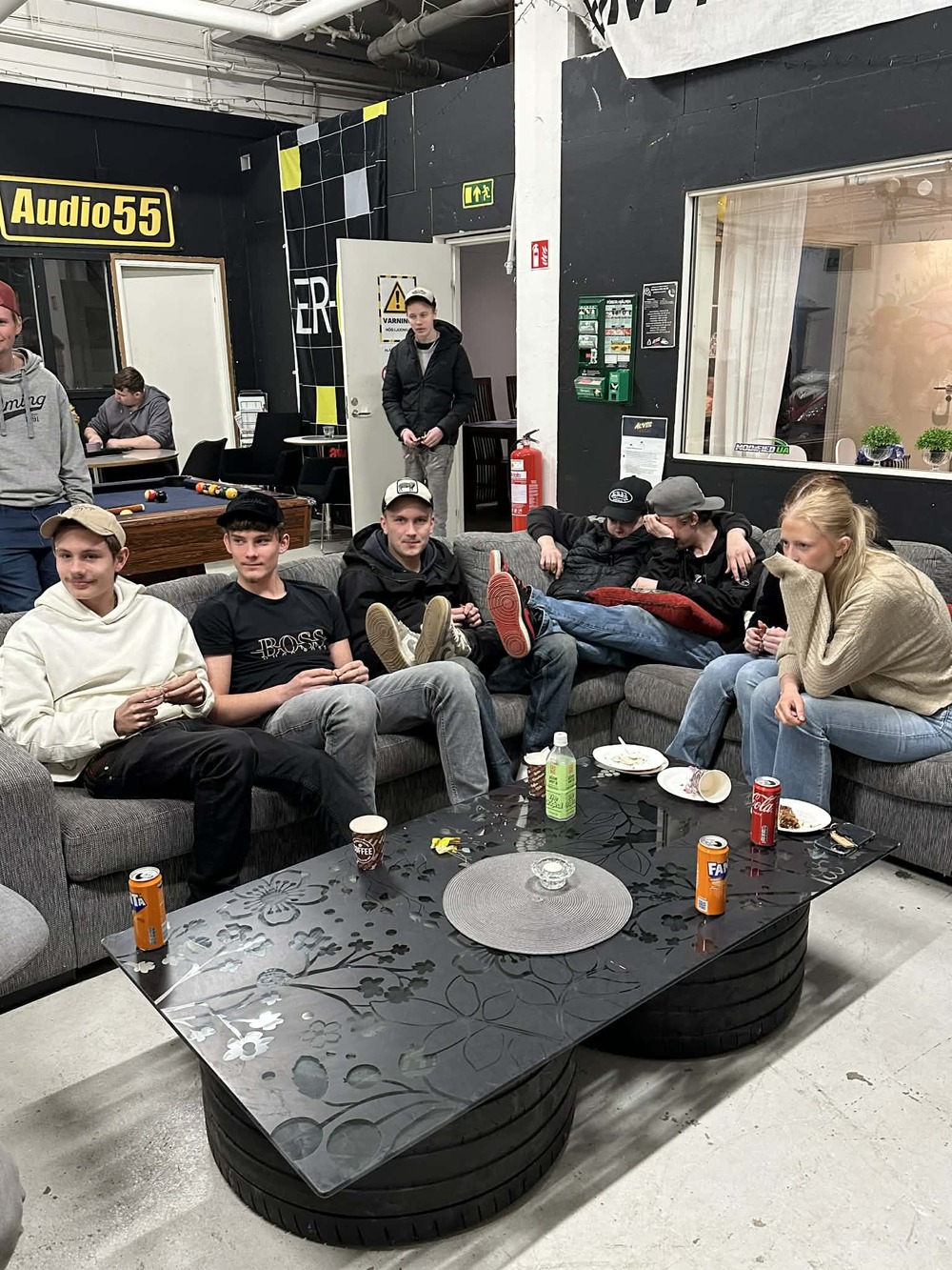 En grupp unga vuxna sitter i en soffa runt ett svart soffbord med drycker och tallrikar i ett vardagsrumsliknande rum.