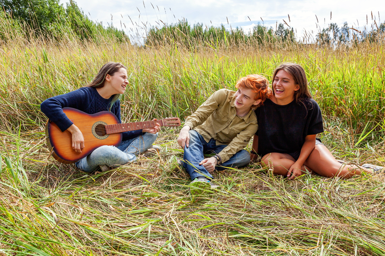 Tre unga personer sitter i gräset, en spelar gitarr och de andra två lyssnar och skrattar.