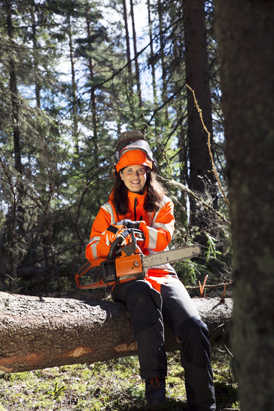 En kvinna i orange skyddskläder sitter på en fälld trädstam i skogen med en motorsåg i händerna.